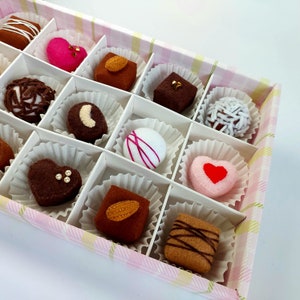 May include: A box of felt chocolates in various shapes and colors, including hearts, swirls, and squares. The chocolates are arranged in a grid pattern and are displayed on a white background.