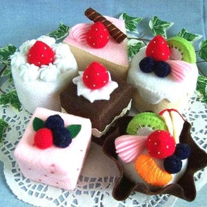 Peut inclure: Six gâteaux en feutre décorés de fruits et de garnitures variés. Les gâteaux sont de formes et de tailles différentes, et ils sont tous très réalistes. Les gâteaux sont disposés sur une dentelle blanche.