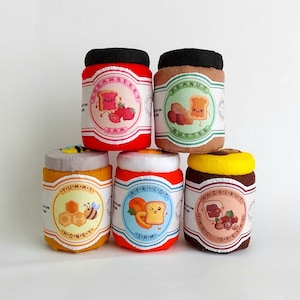 May include: Five colorful fabric jars with lids, each with a different flavor of jam or spread. The jars are labeled "Strawberry Jam", "Peanut Butter", "Honey", "Apricot Jam", and "Hazelnut Spread".