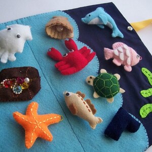 Op de afbeelding: Een vilt activiteit boek met een oceaan thema, met verschillende zeedieren en schatten. Inclusief een witte inktvis, rode krab, oranje zeester, bruine schatkist en een blauwe dolfijn. De achtergrond is blauw en marine met geel lint.