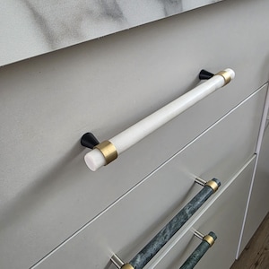 Könnte beinhalten: Nahaufnahme von Schrank Schubladen mit dekorativen Griffen. Die obere Schublade hat einen weißen Griff in Marmoroptik mit goldenen Akzenten und schwarzen Endkappen. Darunter haben zwei Schubladen Griffe in grüner Marmoroptik mit goldenen und silbernen Akzenten. Der Schrank ist hellgrau.