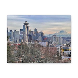 May include: A canvas print featuring a scenic view of Seattle, Washington. The Space Needle is prominent, with the city skyline and a mountain range in the background. The sky is a mix of blue and gray.