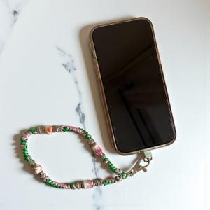 以下が含まれることがあります： 透明なケースに入ったスマートフォンがリストストラップに接続されています。 ストラップは、緑とピンクのコードに花のビーズとシルバーのアクセントが付いています。 電話は白い大理石の表面に置かれています。