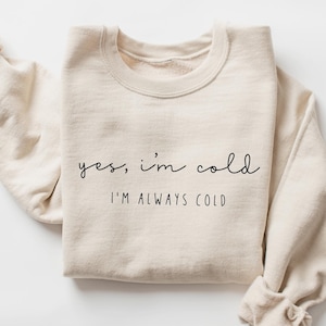 Może przedstawiać: Beżowa bluza z napisem "yes, I'm cold" i "I'M ALWAYS COLD" czarnym skryptem. Długie rękawy są podwinięte. Bluza wykonana jest z miękkiego materiału.