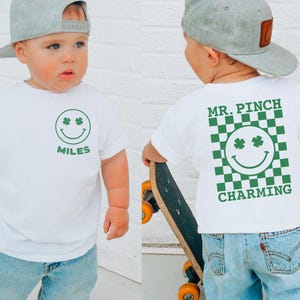 Pode incluir: Uma camiseta branca com um rosto sorridente verde e trevos, com o nome "Miles" impresso abaixo. A parte de trás da camisa apresenta um design xadrez verde e branco com as palavras "Mr. Pinch Charming". A criança está usando um boné cinza e jeans azul claro.