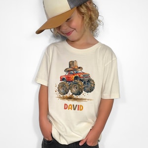 Pode incluir: Uma camiseta creme com uma ilustração em aquarela de um caminhão monstro vermelho com um chapéu de cowboy. O nome "DAVID" está impresso em laranja abaixo. A criança está usando um boné de caminhoneiro com aba marrom e parte superior cinza.