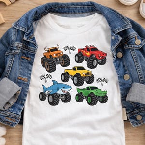 May include: White t-shirt with five cartoon monster trucks: orange with horns, red with flames, yellow, a shark, and a green alligator. Each truck has large black tyres and is set against a checkered flag background. A denim jacket and shoes are in the background.