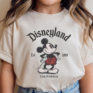 May include: Cream-colored t-shirt featuring a vintage Disneyland graphic. The design includes Mickey Mouse, the text "Disneyland," "Est. 1955," and "California" in black. The shirt has short sleeves and a classic crew neck.