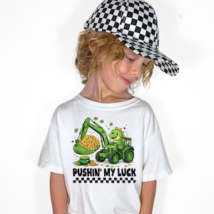 Puede incluir: Una camiseta blanca con un dinosaurio verde operando una excavadora verde, recogiendo monedas de oro en una olla. El texto "PUSHIN' MY LUCK" está debajo del gráfico. La persona lleva una gorra de béisbol a cuadros blancos y negros.