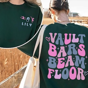 May include: Dark green sweatshirt with the text "LILY" and a gymnast graphic on the front. The back of the sweatshirt features the words "VAULT BARS BEAM FLOOR" in pink, blue, and white, with heart and star accents. A white tote bag is visible.
