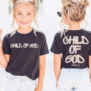 May include: Black t-shirt with the text "CHILD OF GOD" in a light beige color on the front and back. The back of the shirt also includes "PSALM 23". The shirt is paired with light blue denim shorts.
