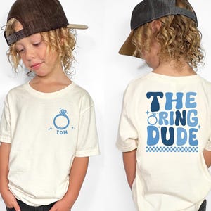 May include: Off-white t-shirt with a blue ring graphic and the name "TOM". The back of the shirt says "THE RING DUDE" in blue lettering. The shirt is worn by a child.