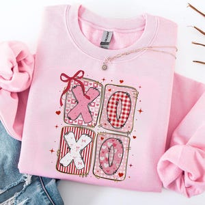 May include: A light pink sweatshirt with a graphic design featuring the letters "XO" in a patchwork style. The letters are in pink, red, and white patterns, with hearts and a red bow. The sweatshirt is made of a soft material.