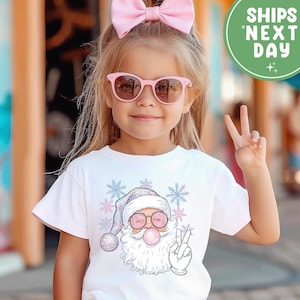 May include: A child wearing a white t-shirt featuring a cartoon Santa Claus blowing a bubblegum bubble and giving a peace sign. The Santa graphic wears sunglasses and a hat. The image also includes the text "SHIPS NEXT DAY".