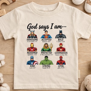 Op de afbeelding: Crèmekleurig T-shirt met de tekst "God says I am..." en illustraties van superhelden. Elke superheld heeft een bijbehorend woord en een bijbelvers. Het T-shirt is omgeven door kinderspeelgoed.
