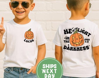 May include: White t-shirt featuring a pumpkin design. The front displays a smiling pumpkin with the name "Lucas". The back of the shirt has a pumpkin graphic and the text "He is the light in the darkness John 1:5".