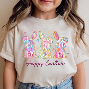 Puede incluir: Una camiseta beige con tres diseños de conejitos de Pascua coloridos, cada uno con un lazo y detalles florales. Los conejitos son en tonos rosa, azul, amarillo y verde. Las palabras "Happy Easter" están escritas en cursiva debajo.