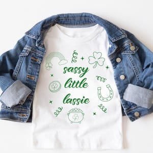 May include: A white t-shirt with the words "sassy little lassie" in green, along with green shamrocks, a rainbow, a horseshoe, and a pot of gold. The shirt is under a blue denim jacket.