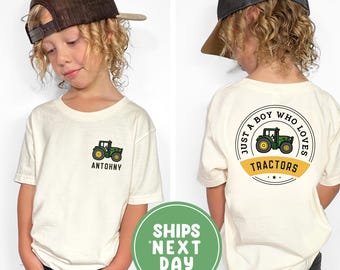 May include: A cream-colored t-shirt featuring a green tractor graphic and the name "ANTOHNY" on the front. The back of the shirt has a circular design with a tractor and the text "JUST A BOY WHO LOVES TRACTORS". The child is wearing a brown cap.