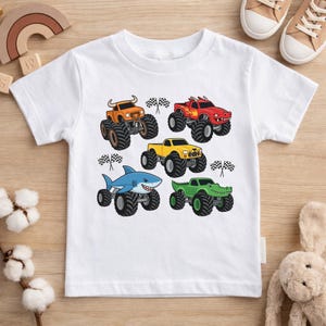 May include: White t-shirt with a design of monster trucks in various colours. The trucks are orange, red, yellow, blue, and green, each with unique details. Checkered flags are also included in the design.