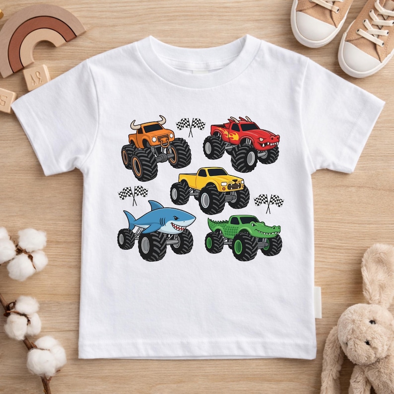May include: White t-shirt with a design of monster trucks in various colours. The trucks are orange, red, yellow, blue, and green, each with unique details. Checkered flags are also included in the design.