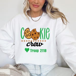 Peut inclure: Sweat-shirt blanc avec l'inscription "COOKIE CREW" en vert et noir, avec un motif de biscuit. En dessous, un c&oelig;ur vert et les mots "Troop 2118". Le sweat-shirt est de style d&eacute;contract&eacute; et confortable.