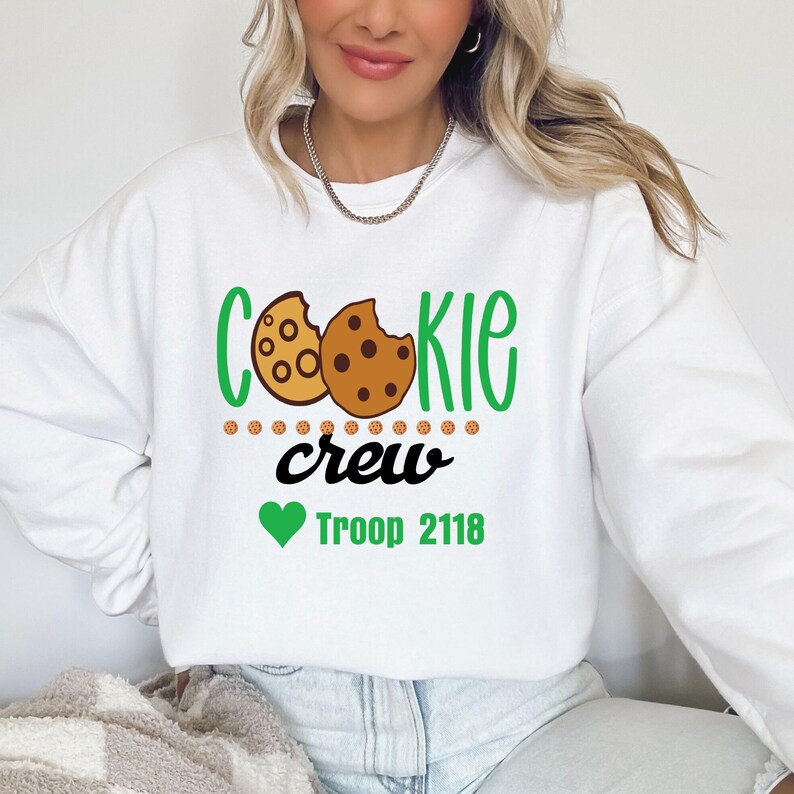 Peut inclure: Sweat-shirt blanc avec l'inscription "COOKIE CREW" en vert et noir, avec un motif de biscuit. En dessous, un c&oelig;ur vert et les mots "Troop 2118". Le sweat-shirt est de style d&eacute;contract&eacute; et confortable.