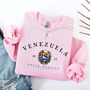 May include: A pale pink sweatshirt featuring the word "VENEZUELA" in black, arched lettering. Beneath the word is a coat of arms and the numbers "18" and "11". The words "SOUTH AMERICA" and coordinates are printed below the coat of arms. A necklace is draped across the sweatshirt.