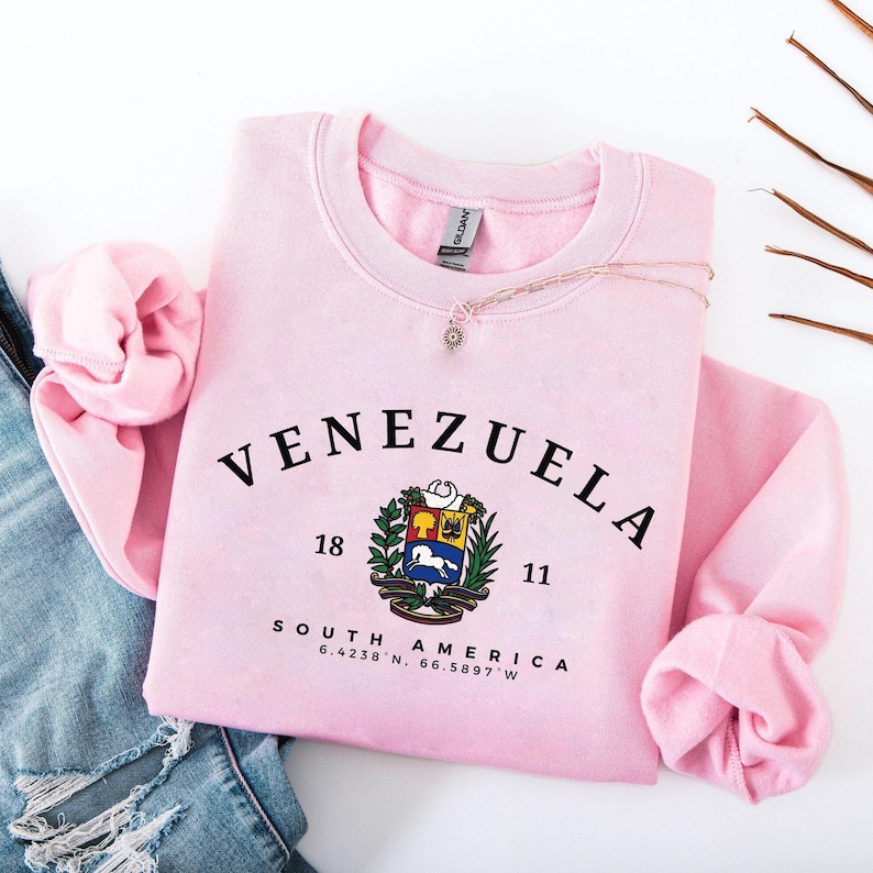 May include: A pale pink sweatshirt featuring the word "VENEZUELA" in black, arched lettering. Beneath the word is a coat of arms and the numbers "18" and "11". The words "SOUTH AMERICA" and coordinates are printed below the coat of arms. A necklace is draped across the sweatshirt.