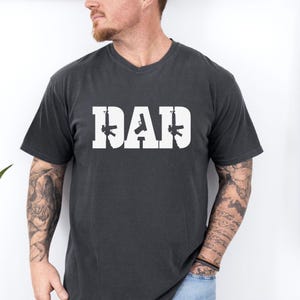 May include: Dark gray t-shirt with the word "DAD" in white block letters. The letter "A" is replaced with a handgun, and the letters "D" are replaced with rifles. The shirt has a V-neck and short sleeves.