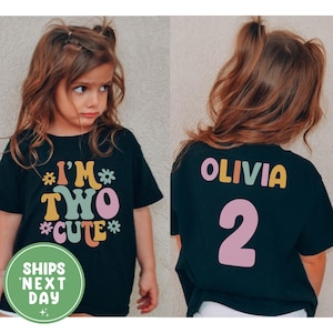 Op de afbeelding: Zwart T-shirt met de tekst "I'M TWO CUTE" in kleurrijke letters en bloemen. De achterkant van het shirt heeft de naam "OLIVIA" en het nummer "2". Het shirt is voor een kind.