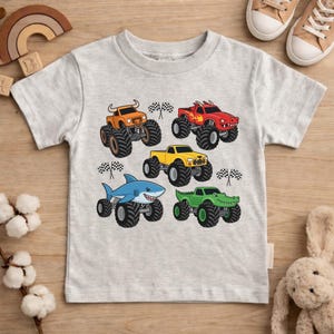 May include: A light grey t-shirt with five colourful monster truck designs. The trucks are orange, red, yellow, blue, and green, each with unique monster truck features. Checkered flags are also included in the design.