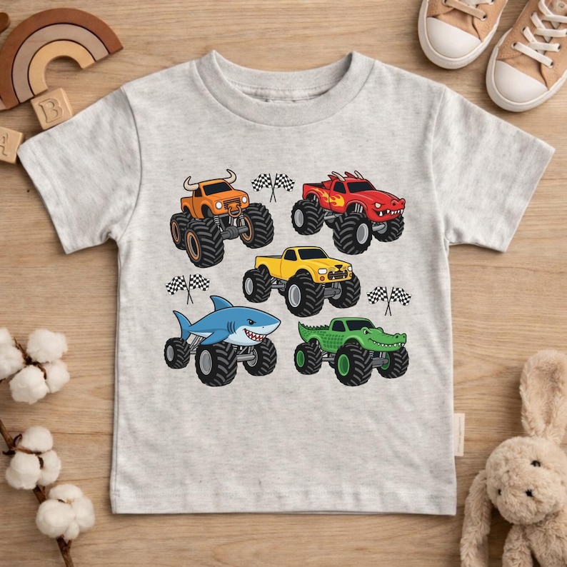 May include: A light grey t-shirt with five colourful monster truck designs. The trucks are orange, red, yellow, blue, and green, each with unique monster truck features. Checkered flags are also included in the design.