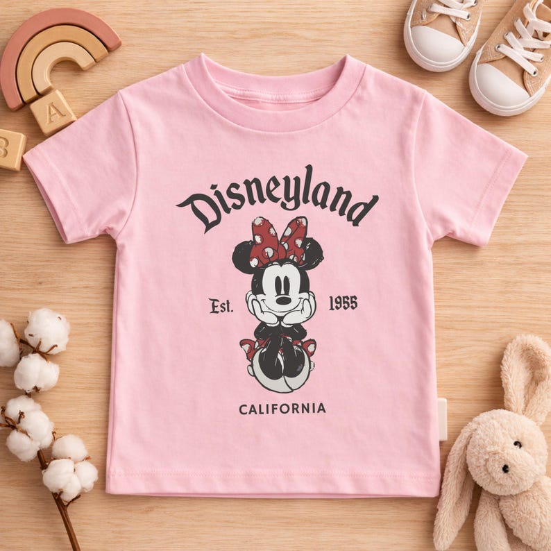 May include: Pink t-shirt featuring a Minnie Mouse graphic with a red bow and the text "Disneyland Est. 1955 California". The shirt is surrounded by children's toys, including a wooden rainbow and a stuffed bunny.