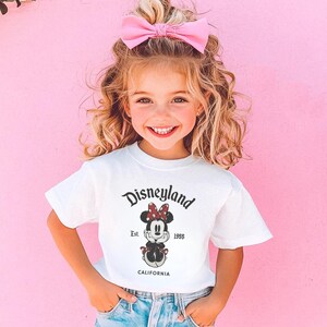 May include: A young girl wearing a white t-shirt with a Disneyland graphic featuring Minnie Mouse. The shirt has the text "Disneyland" above the Minnie Mouse graphic, with "Est. 1955" and "CALIFORNIA" below. She is wearing a pink bow and blue jeans.