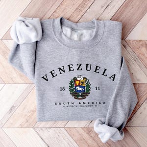 May include: A grey sweatshirt featuring the word "VENEZUELA" in black arched text. Below is a colourful emblem and the words "SOUTH AMERICA" with coordinates. The sleeves are folded on a wooden surface.