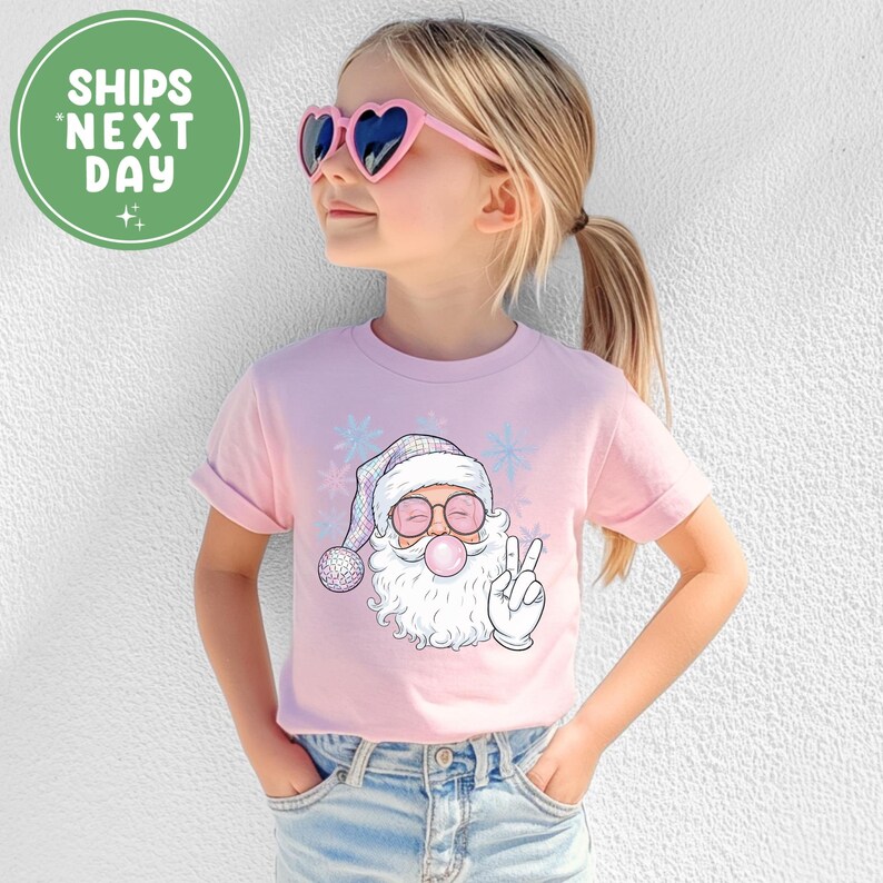 May include: A light pink t-shirt with a cartoon Santa Claus blowing a bubblegum bubble and giving a peace sign. The Santa wears a hat and glasses, and has a white beard. The shirt has the text "SHIPS NEXT DAY" in a green circle.