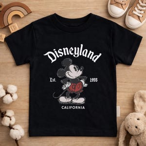May include: Black t-shirt featuring a graphic of Mickey Mouse with the text "Disneyland" above and "Est. 1955 CALIFORNIA" below. The cartoon Mickey Mouse is in a standing pose, wearing red shorts and white gloves.