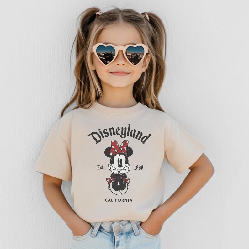 May include: A young person wearing heart-shaped sunglasses and a beige t-shirt. The t-shirt features the Disneyland logo and a Minnie Mouse graphic. The text "Est. 1955 CALIFORNIA" is printed below the graphic.