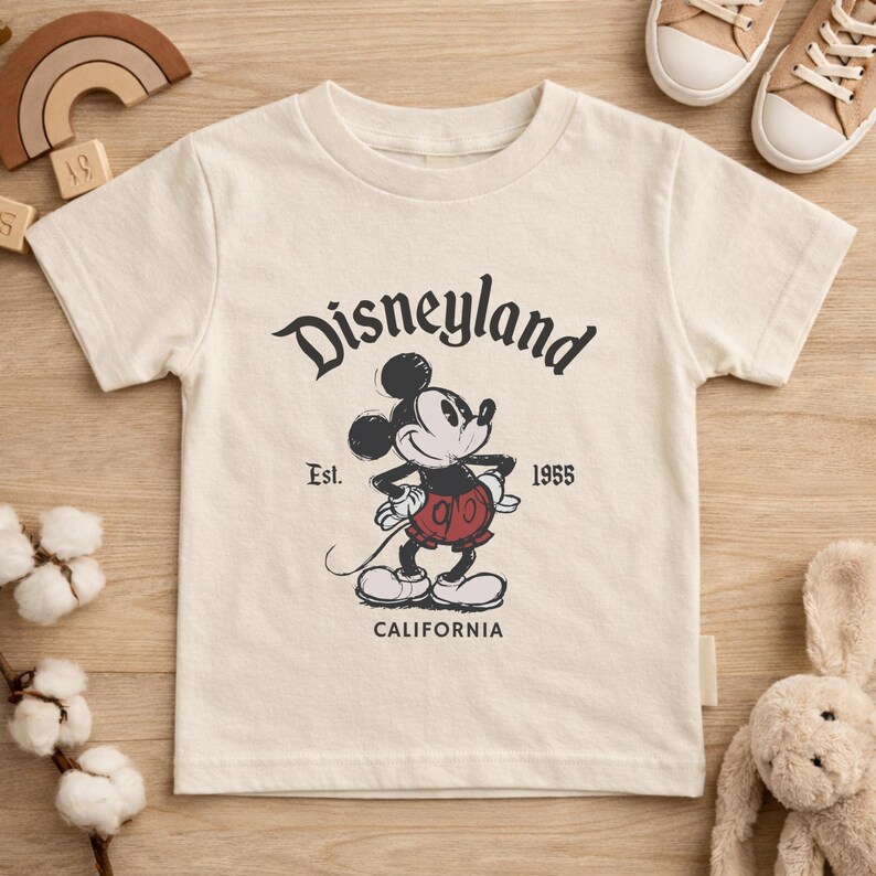 May include: Cream-colored t-shirt featuring a classic Mickey Mouse graphic with the text "Disneyland" above the character. The shirt also includes "Est. 1955" and "CALIFORNIA" in black lettering. The shirt is surrounded by other toys.