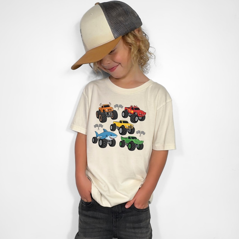 May include: A cream-coloured t-shirt with six colourful monster truck designs. The trucks are orange, red, yellow, green, and a shark-themed blue truck. Each truck has large tyres and is set against a chequered flag background.