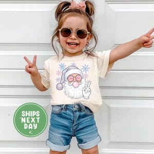 May include: A cream-coloured t-shirt featuring a cartoon Santa Claus blowing a bubblegum bubble, wearing sunglasses and a peace sign. The Santa graphic is surrounded by snowflakes. The child is wearing denim shorts and sunglasses.