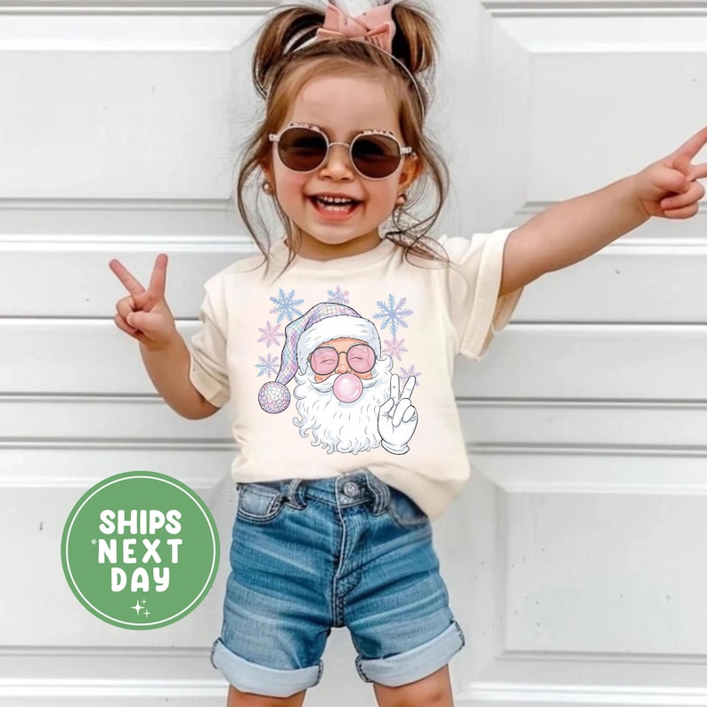 May include: A cream-coloured t-shirt featuring a cartoon Santa Claus blowing a bubblegum bubble, wearing sunglasses and a peace sign. The Santa graphic is surrounded by snowflakes. The child is wearing denim shorts and sunglasses.