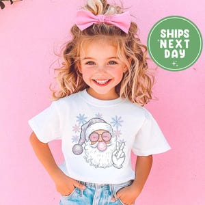 May include: A white t-shirt featuring a cartoon Santa Claus with sunglasses blowing a bubblegum bubble and making a peace sign. The Santa Claus is wearing a plaid hat and surrounded by snowflakes.