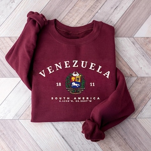 May include: A maroon sweatshirt featuring the word "VENEZUELA" in white arched lettering. Below is a colourful emblem and the words "SOUTH AMERICA" with coordinates. The sweatshirt has long sleeves and is folded on a wooden surface.