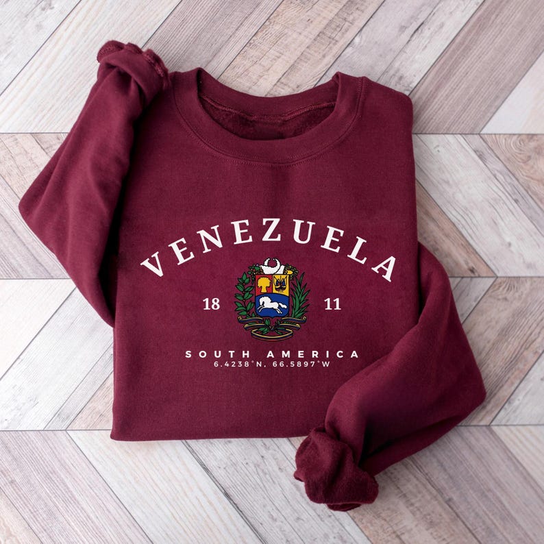 May include: A maroon sweatshirt featuring the word "VENEZUELA" in white arched lettering. Below is a colourful emblem and the words "SOUTH AMERICA" with coordinates. The sweatshirt has long sleeves and is folded on a wooden surface.
