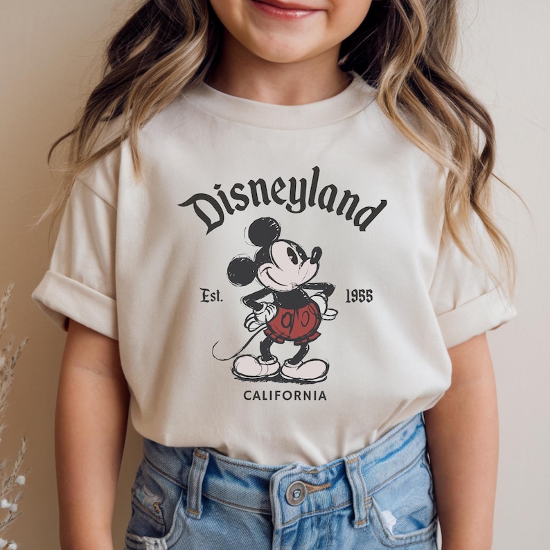May include: Cream-colored t-shirt featuring a vintage Disneyland graphic. The design includes Mickey Mouse, the text "Disneyland," "Est. 1955," and "California" in black. The shirt has short sleeves and a classic crew neck.