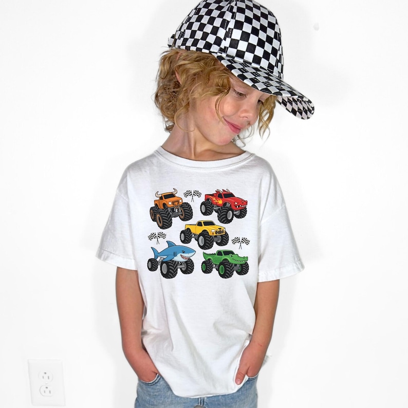 May include: A white t-shirt with colourful monster truck graphics. The shirt shows six different monster truck designs in various colours, including orange, red, yellow, green, and a shark-themed truck. The child is wearing a black and white chequered baseball cap.