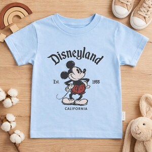 May include: A light blue t-shirt featuring a vintage-style graphic of Mickey Mouse with the text "Disneyland" above and "Est. 1955 California" below. The shirt is a short-sleeved design.