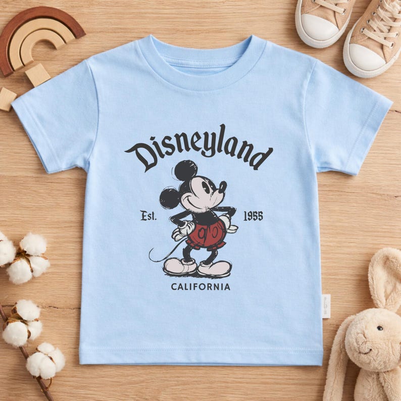 May include: A light blue t-shirt featuring a vintage-style graphic of Mickey Mouse with the text "Disneyland" above and "Est. 1955 California" below. The shirt is a short-sleeved design.
